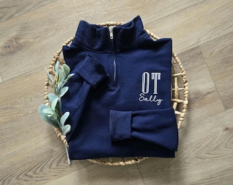 May include: A navy blue quarter-zip sweatshirt with the letters "OT" and the name "Sally" embroidered in white. The sweatshirt is folded and placed in a woven basket with a sprig of greenery.