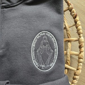 Puede incluir: Una sudadera con capucha gris carb&oacute;n que presenta un parche ovalado bordado en blanco. El parche muestra una figura con los brazos extendidos, rodeada por el texto: O MARY, CONCEIVED WITHOUT SIN, PRAY FOR US WHO HAVE RECOURSE TO THEE.