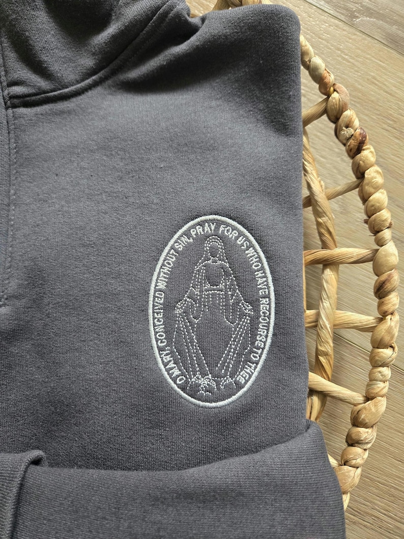 Puede incluir: Una sudadera con capucha gris carb&oacute;n que presenta un parche ovalado bordado en blanco. El parche muestra una figura con los brazos extendidos, rodeada por el texto: O MARY, CONCEIVED WITHOUT SIN, PRAY FOR US WHO HAVE RECOURSE TO THEE.