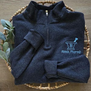 May include: A dark gray quarter-zip sweatshirt with a blue embroidered pharmacy symbol and the text "Alexis, PharmD".