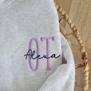 May include: A grey sweatshirt with the letters "OT" embroidered in purple and the name "Alexa" written in black cursive.
