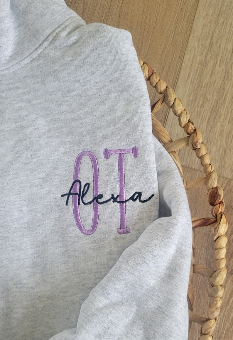 May include: A grey sweatshirt with the letters "OT" embroidered in purple and the name "Alexa" written in black cursive.