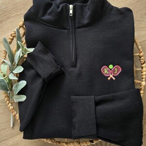 May include: Black quarter-zip sweatshirt with a pink and green embroidered design of two tennis rackets and a tennis ball. The rackets have the letters "K" and "J" embroidered on them.
