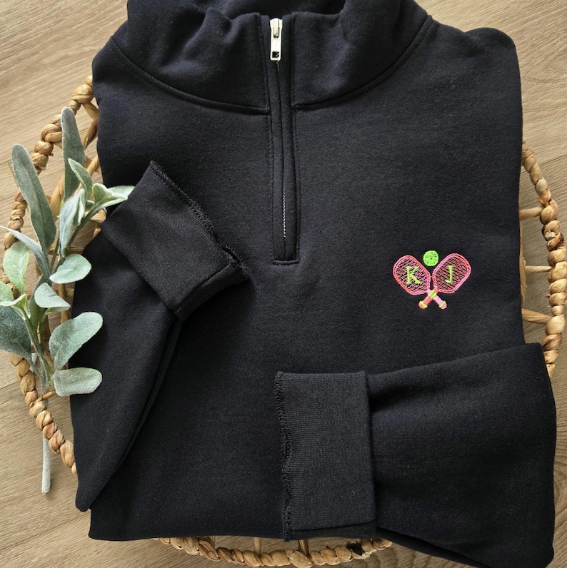 May include: Black quarter-zip sweatshirt with a pink and green embroidered design of two tennis rackets and a tennis ball. The rackets have the letters "K" and "J" embroidered on them.