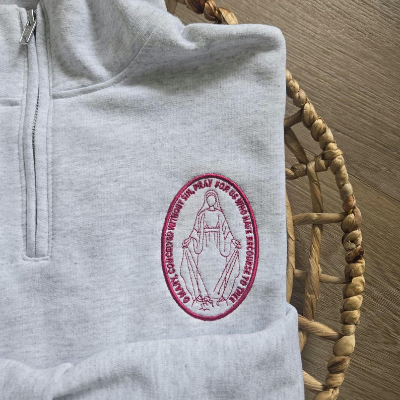 Puede incluir: Una sudadera gris claro con cremallera de cuarto y un parche ovalado bordado en el pecho izquierdo. El bordado rosa representa una figura con t&uacute;nica rodeada por el texto O MARY, CONCEIVED WITHOUT SIN, PRAY FOR US WHO HAVE RECOURSE TO THEE.
