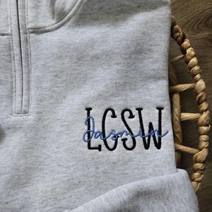 May include: A grey quarter-zip sweatshirt with the name "Jasmine" embroidered in blue and black lettering.