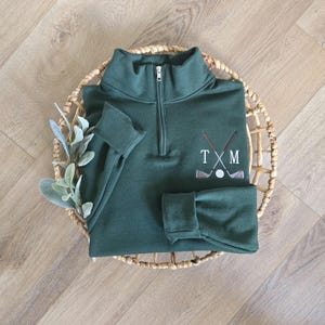 May include: A dark green golf pullover with a quarter-zip neckline and long sleeves. The pullover features an embroidered design of crossed golf clubs, a golf ball, and the letters "T X M". The pullover is displayed on a woven basket.