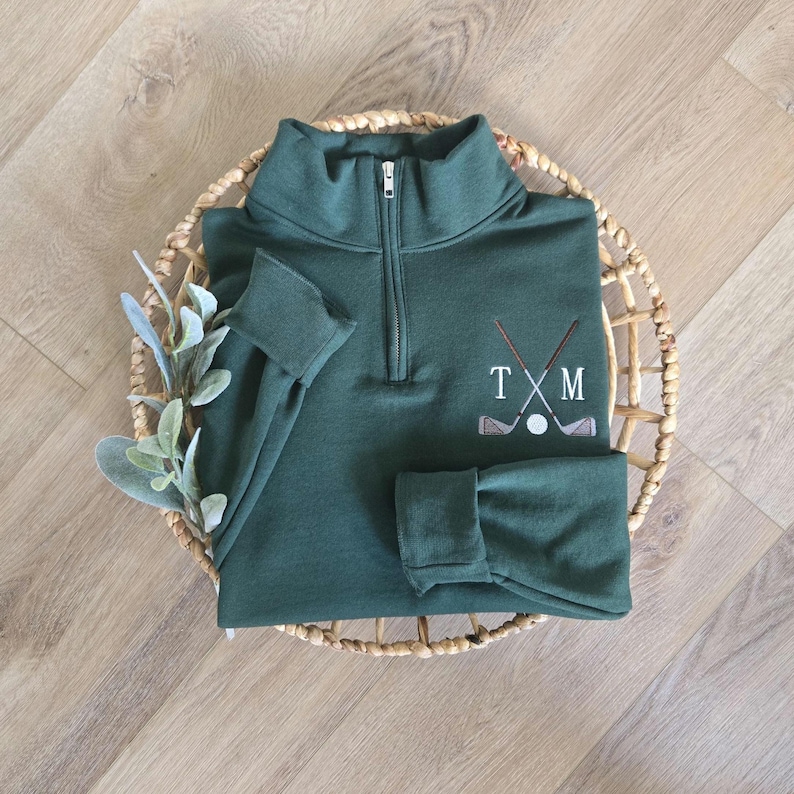 Peut inclure: Un pull de golf vert fonc&eacute; avec un col montant zipp&eacute; et des manches longues. Le pull pr&eacute;sente un motif brod&eacute; de clubs de golf crois&eacute;s, d'une balle de golf et des lettres "T X M". Le pull est pr&eacute;sent&eacute; sur un panier tress&eacute;.