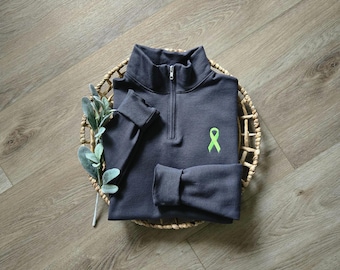 Può includere: Una felpa grigio antracite con zip a un quarto e un nastro di sensibilizzazione verde neon ricamato sul petto. La felpa è piegata in un cestino intrecciato con un rametto di verde.