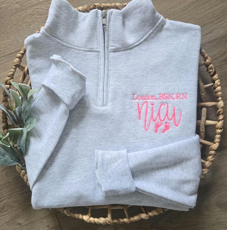 K&ouml;nnte beinhalten: Hellgraues Sweatshirt mit Viertelrei&szlig;verschluss und dem Text "London, BSN, RN" &uuml;ber dem Namen "Nia" in rosa Stickerei. Das Design beinhaltet rosa Fu&szlig;abdr&uuml;cke. Das Sweatshirt ist in einem geflochtenen Korb gefaltet.