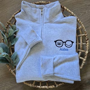 May include: A light gray quarter-zip sweatshirt with a black glasses and eye design and the name "Aidan" in blue. The sweatshirt is folded in a woven basket.