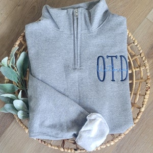 May include: A grey quarter-zip sweatshirt with a blue embroidered monogram that says "OTD".