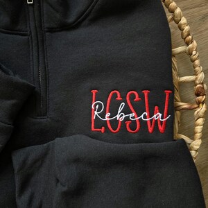 May include: Black quarter-zip sweatshirt with red embroidered lettering that says "LCSW Rebecca".