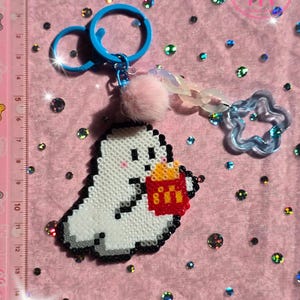 Könnte beinhalten: Ein weißer Geister-Schlüsselanhänger aus Pixelperlen, der einen roten Behälter mit gelben Pommes hält. Er verfügt über einen blauen Kunststoffring, einen kleinen rosa Pompon und einen durchsichtigen blauen Sternanhänger. Ein Lineal zeigt 9 cm Höhe.