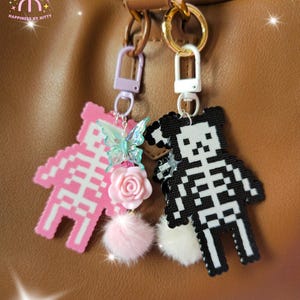 Op de afbeelding: Twee sleutelhangers in pixel-artstijl in de vorm van skeletberen aan een bruine tas. De ene is roze en wit met een doorschijnende vlinder, een roze roos en een roze pompon. De andere is zwart en wit met een witte pompon. Beide hebben metalen sluitingen.