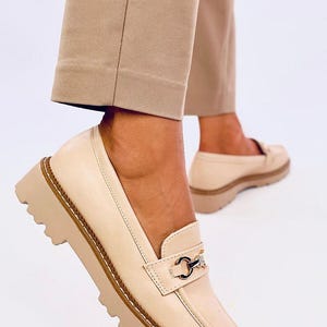 May include: Beige leather loafers with a chunky platform sole and a gold chain detail. The loafers have a pointed toe and a slip-on design.