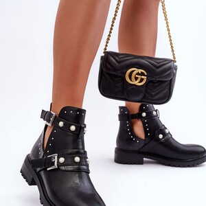 May include: Black ankle boots with cut-out sides, pearl detailing, and buckle fastenings. The boots feature a chunky sole. A black quilted crossbody bag with a gold chain strap and a gold logo is also visible.