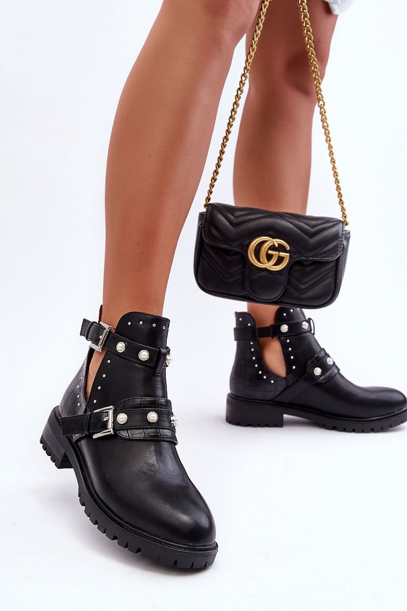 May include: Black ankle boots with cut-out sides, pearl detailing, and buckle fastenings. The boots feature a chunky sole. A black quilted crossbody bag with a gold chain strap and a gold logo is also visible.