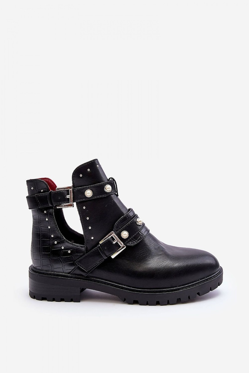 May include: Black ankle boot with a cut-out design, featuring multiple straps with silver buckles and faux pearl accents. The boot has a textured, crocodile-like pattern on the side and a lug sole.