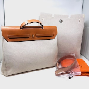 May include: A structured handbag featuring a beige canvas body and a tan leather flap with a top handle. The bag includes a matching beige canvas pouch with a central snap closure, a tan leather shoulder strap in a plastic bag, and an orange fabric pouch.