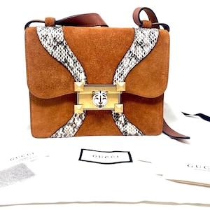 May include: A brown suede shoulder bag featuring snakeskin-patterned leather accents, a gold-tone metal clasp with a tiger head emblem, and four pyramid studs. The bag is displayed with white dust bags featuring the Gucci logo in black text.