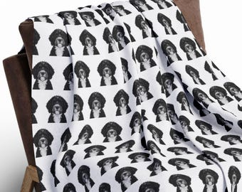 May include: A white throw blanket draped over a wooden chair. The blanket is covered in a repeating pattern of a black and white image of a dog wearing a hat. The blanket is folded over the chair and extends to the floor.