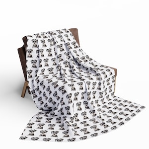 Peut inclure: Une couverture en polaire blanche avec un motif répété de la tête d'un petit chien gris. La couverture est pliée et drapée sur un fauteuil marron.