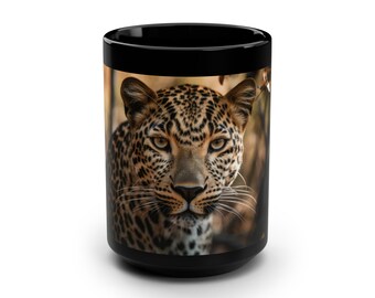 May include: A black ceramic mug featuring a high-resolution photographic print of a leopard's face. The leopard has golden eyes, black spots, and white whiskers, centered on the front of the mug against a blurred, natural brown background.