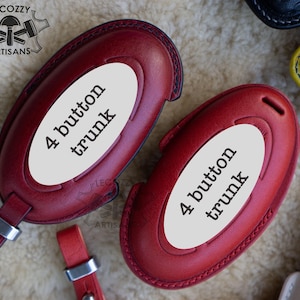 May include: Two red leather key fob covers with the text "4 button trunk" on an oval label. One cover has a key ring and a red strap with a silver clasp. The covers are on a textured surface.