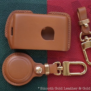 May include: A set of two keyrings made of brown leather and gold metal. The keyrings feature a round leather fob and a rectangular leather case with a cut-out for a key. The keyrings are labeled "Lecozzy Artisans".