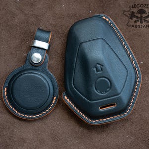 May include: Black leather key fob cover and round AirTag holder with orange stitching. The key fob cover has an arrow symbol and a circular indentation. The AirTag holder has a silver metal clasp. The logo "LECOZZY ARTISANS" is in the upper right corner.
