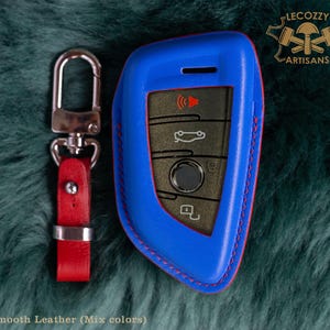 May include: Blue leather key fob with red stitching. The fob has a silver key ring attached. The fob has a logo that says "Lecozzy Artisans".