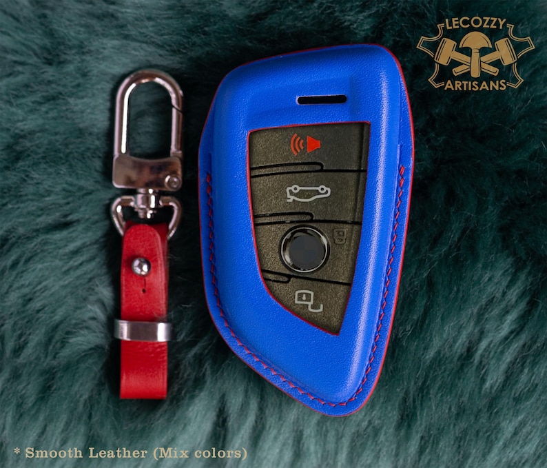 May include: Blue leather key fob with red stitching. The fob has a silver key ring attached. The fob has a logo that says "Lecozzy Artisans".