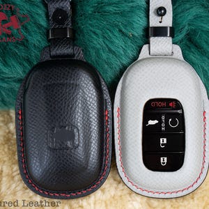 May include: Two leather key fobs, one black and one gray, with red stitching. The gray fob has a car key with buttons labeled "Hold" and "Unlock".