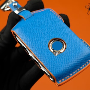 May include: A blue leather key fob with an orange stitch detail and a silver button. The fob is designed to protect a car key.