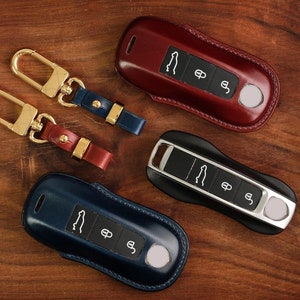 Puede incluir: Tres fundas de cuero para llaves en rojo, azul y negro. Cada funda tiene un botón plateado y un llavero plateado con un logotipo de coche. Las fundas están sobre una superficie de madera.