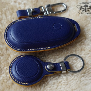 May include: A set of blue leather keychains and key fob cases with yellow stitching. The keychains have silver-coloured metal hardware. The leather has a textured appearance. The logo "LECOZZY ARTISANS" is in the upper right corner.