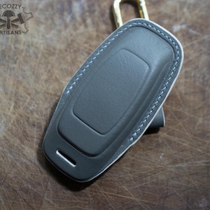 May include: A gray leather key fob cover with white stitching and a gold-colored clasp. The cover has a rectangular shape with a curved top and a small slot on the bottom. The logo "LECOZZY ARTISANS" is visible in the top left corner.