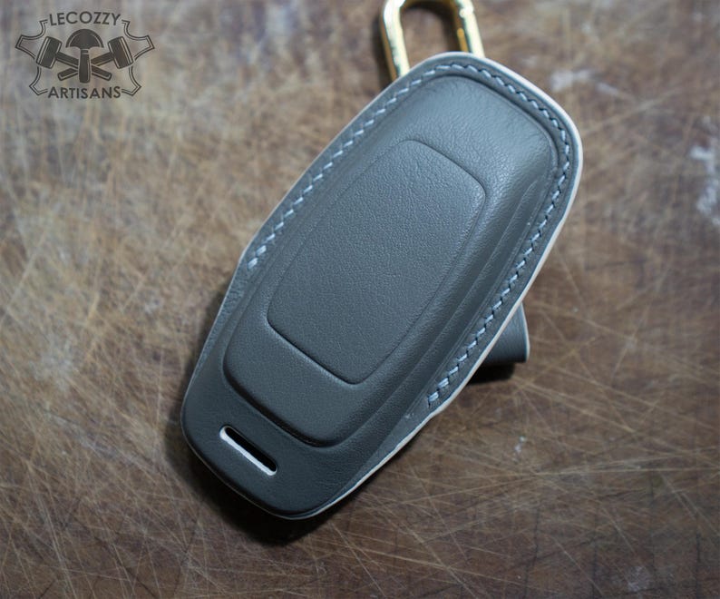 May include: A gray leather key fob cover with white stitching and a gold-colored clasp. The cover has a rectangular shape with a curved top and a small slot on the bottom. The logo "LECOZZY ARTISANS" is visible in the top left corner.