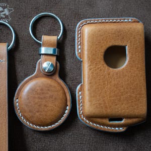 May include: A collection of handcrafted leather accessories in shades of brown. Includes a key fob with a round AirTag holder, a key ring with a leather strap, and a protective case. All items feature white stitching and metal hardware.