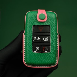Puede incluir: Llavero de cuero verde con costuras rosas y un botón verde. El llavero tiene una llave negra con botones para bloquear, desbloquear, maletero y pánico.
