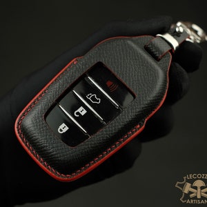May include: Black leather key fob with red stitching and a silver key ring. The fob has four buttons with icons for unlock, lock, boot, and panic.