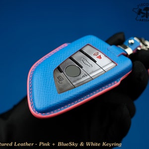 May include: A blue leather key fob with pink stitching and a white keyring. The key fob has a silver car key with buttons for locking, unlocking, and boot release. The text "Lecozzy Artisans" is visible in the top right corner of the image.