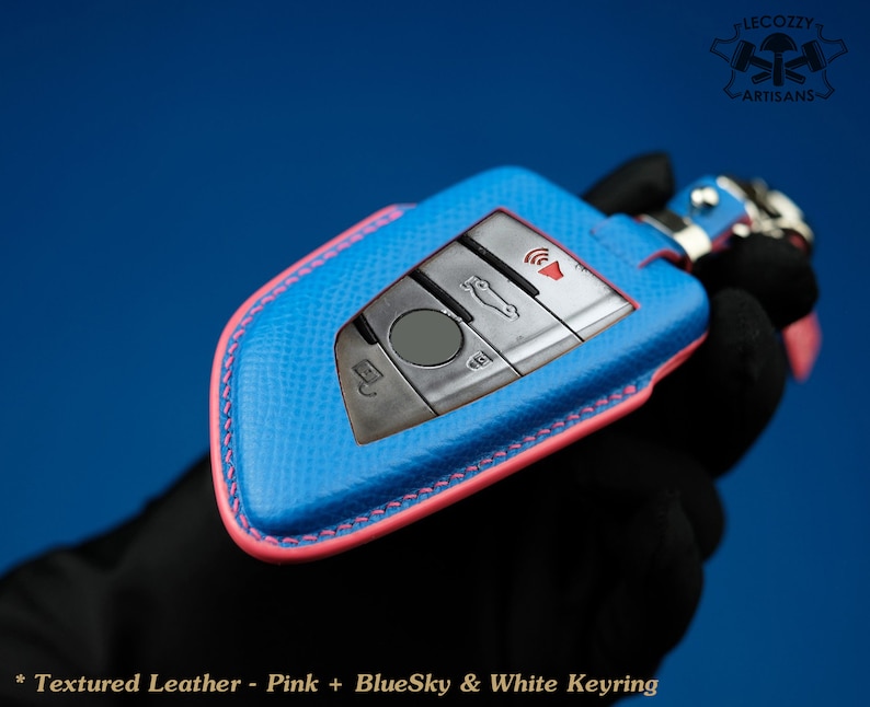 May include: A blue leather key fob with pink stitching and a white keyring. The key fob has a silver car key with buttons for locking, unlocking, and boot release. The text "Lecozzy Artisans" is visible in the top right corner of the image.