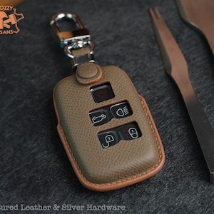 May include: A brown leather key fob with a silver key ring and a textured surface. The fob has five buttons with symbols for a car, headlights, and a lock. The text "*Textured Leather & Silver Hardware" is visible at the bottom of the image.
