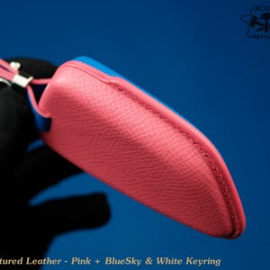 May include: A pink leather key chain with a blue leather trim. The keychain has a white keyring and the text "BlueSky & White Keyring" is printed on the bottom.