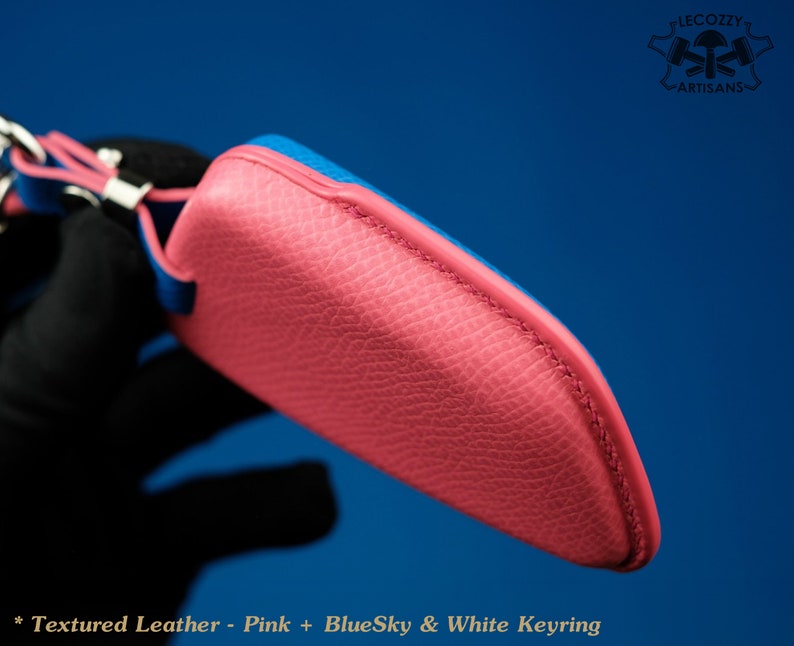 May include: A pink leather key chain with a blue leather trim. The keychain has a white keyring and the text "BlueSky & White Keyring" is printed on the bottom.
