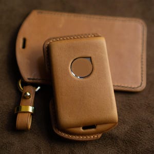 May include: A collection of tan leather accessories, featuring a phone case with a silver detail, a luggage tag, and a keyring with a gold-coloured ring. The items are displayed on a brown textured surface.