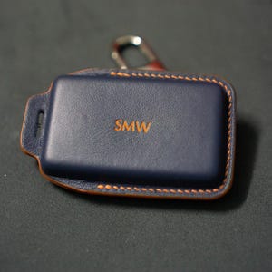 May include: A navy blue leather key fob case with the initials "SMW" in orange. The case has orange stitching around the edges and a silver-coloured metal clasp.