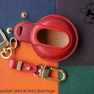 Può includere: Portachiavi in ​​pelle rossa con un anello in metallo dorato e una piccola borsa in pelle rossa. La borsa ha una chiusura a scatto in metallo dorato. Il portachiavi è su una superficie arancione testurizzata.